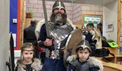 A person dressed as a viking warrior stands with two children, also in viking attire, possibly at a historical or cultural event.