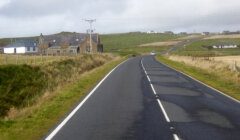 An empty country road leading towards a small collection of buildings with hills in the distance under a cloudy sky.