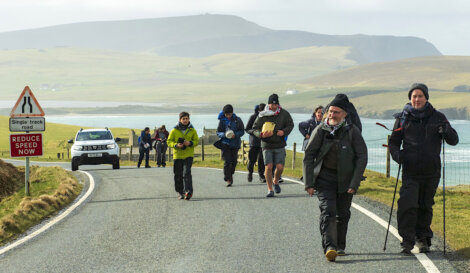Group of hikers walking along a single-track road near a coastal area with a warning sign advising to reduce speed now.