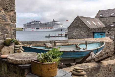 Old wooden boat and stacked stones in the foreground with a large cruise ship anchored in the bay beyond, viewed from a coastal village.