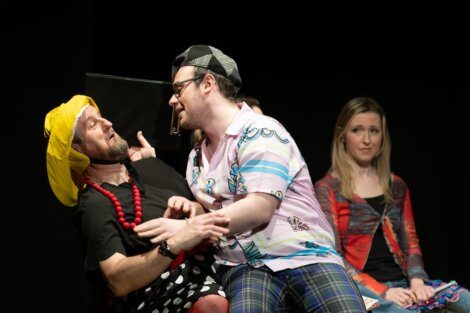 Three actors on stage, with one dressed in colorful, mismatched clothes speaking closely to another in a yellow headwrap, while a third observes with a concerned expression.