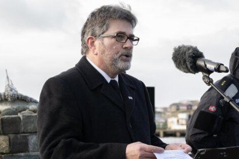A man wearing glasses and a coat reads from a paper at an outdoor event with a microphone in front of him.