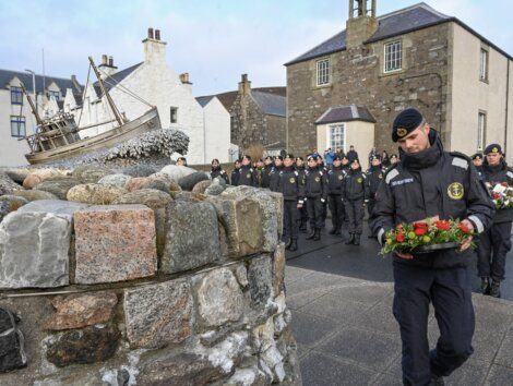 A uniformed individual laying a bouquet of flowers with a group of solemn onlookers in the background.
