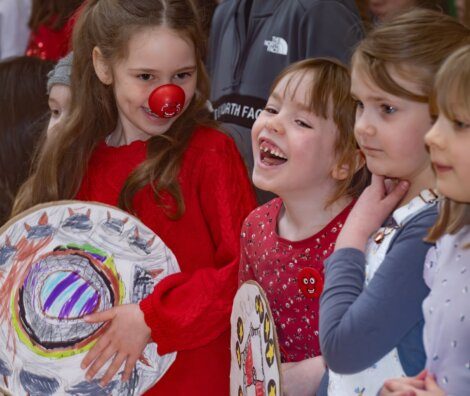 Child with a red nose holding artwork surrounded by other children.