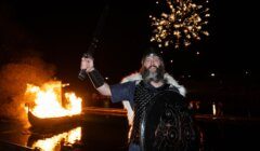 Man in viking costume holding a sword and shield with a burning boat and fireworks in the background.