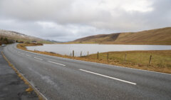 Curving road alongside a calm lake with gentle hills under a cloudy sky.