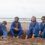 Four men in coveralls sitting on a sandy beach.