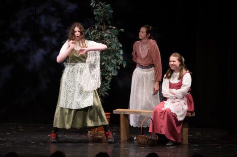 Three actors on stage during a theatrical performance, with one gesturing expressively while the other two watch.