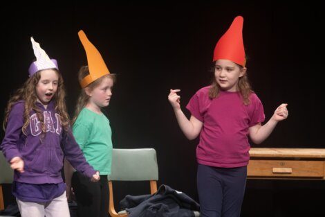 Three children performing on stage, with one wearing a whimsical pointed hat.