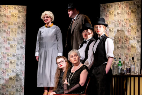 Performers on stage in a theatrical production, set against a backdrop of newspaper clippings, with varied expressions and costumes suggesting character diversity.