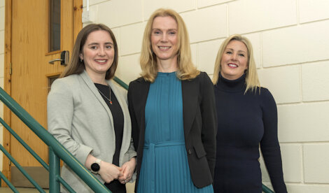 Three women smiling and posing together indoors next to a staircase.
