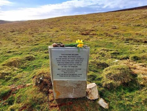 A memorial stone with an inscription and flowers on top sits in a grassy field under a cloudy sky.