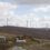 A rural landscape featuring a small road, two houses, and a large wind farm with multiple turbines on a hilly terrain under a cloudy sky.