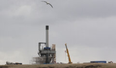 A seagull flies over an industrial construction site with large machinery, cranes, and a tall chimney under a cloudy sky.
