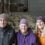 Three generations of a family—a senior man, senior woman, and younger woman—smiling in a barn with sheep in the background.