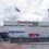 Front view of aberdeen international airport building with two scottish flags flying and a shuttle bus outside.
