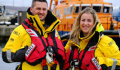 Two lifeboat crew members in yellow rnli jackets smiling in front of a lifeboat.