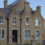 A stone civic building with a pointed roof, labeled "SOUTH RONALDSAY TOWN HALL," featuring chimneys, multiple windows, and a flagpole nearby, situated in a coastal town.