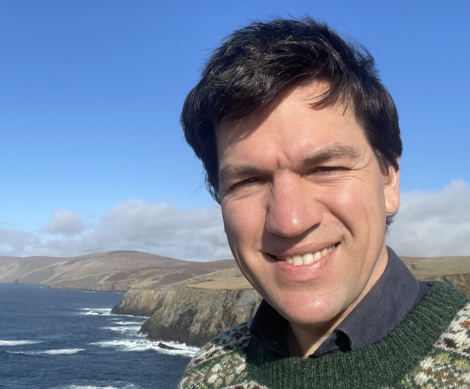 A person smiling for a selfie with a scenic coastal background on a sunny day.
