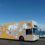 An art bus decorated with black and white illustrations, parked beside a seaside road under a clear blue sky.
