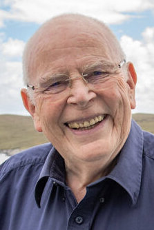 Elderly man with glasses smiling outdoors, wearing a blue shirt against a cloudy sky backdrop.