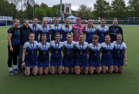 Women's field hockey team posing with medals on a sports field, wearing blue uniforms and smiles, with a coach on the left.
