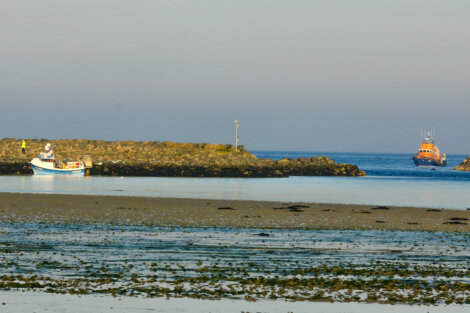Two boats near a rocky breakwater in calm waters with seaweed visible at low tide and a clear sky.