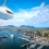 A large white airship with "AirLines" written on it flies over a coastal town with a marina and green hills in the background.