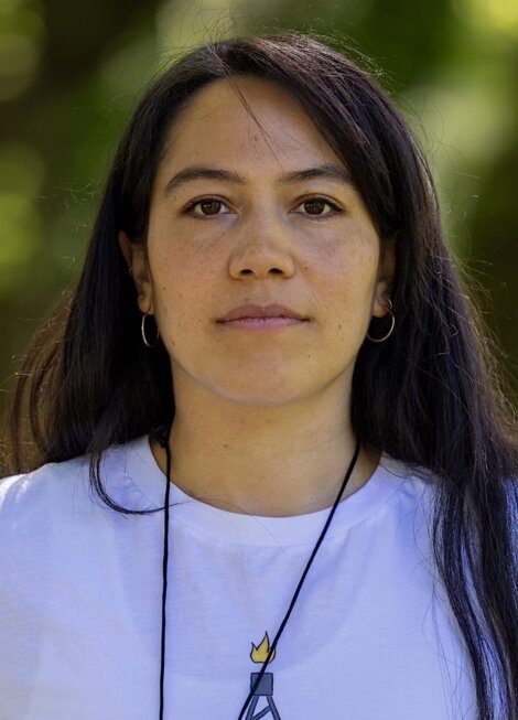 A person with long dark hair, wearing a white shirt and a necklace with a pendant, is looking directly at the camera with a neutral expression. The background is blurred greenery.