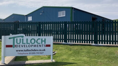 A green and white "Tulloch Developments" sign in front of a blue industrial building with a green fence under a clear sky.