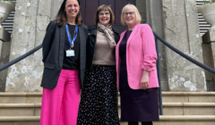 Three women stand on stairs in front of a building. The woman on the left wears a black blazer and pink pants, the woman in the middle wears a black and white polka dot skirt, and the woman on the right wears a pink blazer.
