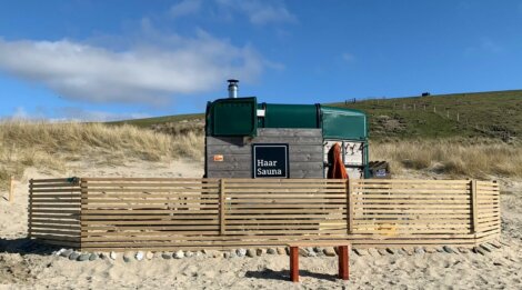 A portable wooden sauna named "Haar Sauna" with a green roof sits on sandy ground, surrounded by a wooden fence. Rolling grassy hills and a clear blue sky are in the background.