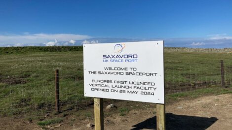 Sign at the SaxaVord UK Space Port reading: "Welcome to the SaxaVord Spaceport - Europe's first licensed vertical launch facility opened on 29 May 2024," against a backdrop of grass and clear sky.