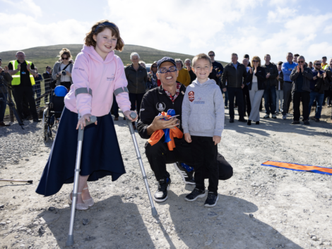 A child using crutches, a man, and another child smile and pose at an outdoor event. An audience stands in the background. A ribbon and pair of scissors are visible.