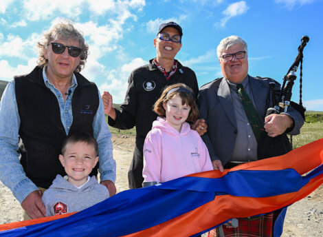 Five people standing outdoors behind a vibrant red, orange, and blue ribbon, with one person holding bagpipes. Two children are also present, along with sunny weather in the background.