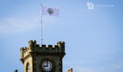 A clock tower with a flag displaying an emblem flies above Lerwick Town Hall. The sky is clear and blue.