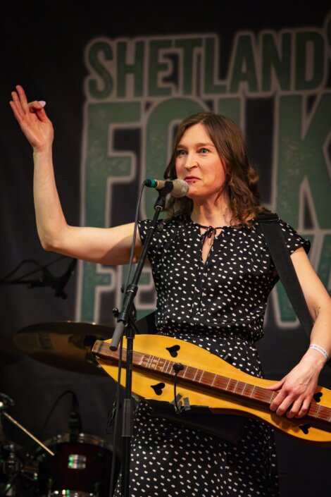 A woman performs at shetland folk festival, singing into a microphone and playing an electric bass, raising one hand in gesture.