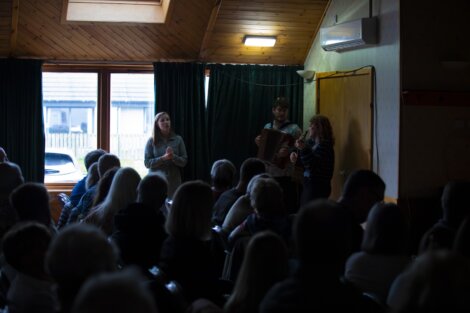 Two musicians playing accordion and singing to an audience in a dimly lit, wood-paneled room.