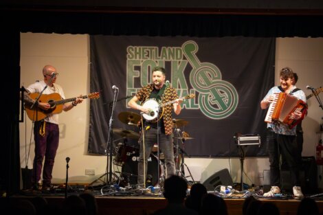 Three musicians performing at the shetland folk festival, featuring a guitarist, a drummer, and an accordion player on stage.
