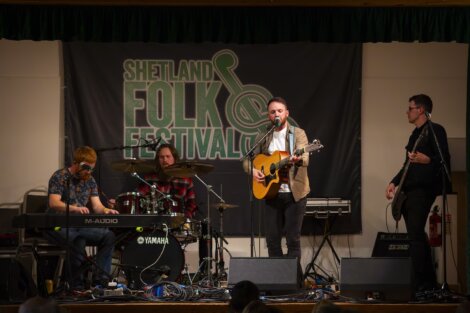 Four musicians performing on stage at the shetland folk festival; drummer, keyboardist, guitarist/vocalist, and bassist with event banner in the background.