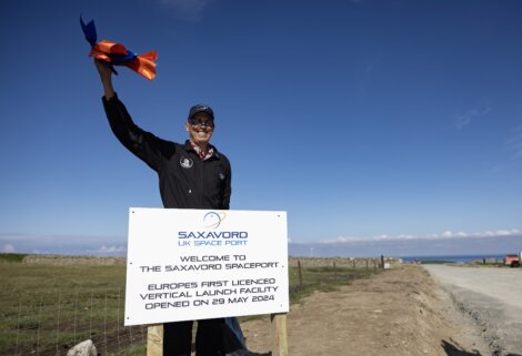 A person stands next to a sign at Saxavord Spaceport, UK's first licensed vertical launch facility, with a rocket model in hand. The sign announces the facility's opening on May 29, 2024.