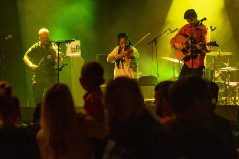 Three musicians perform on stage in front of an audience, featuring a violinist, a guitarist, and another guitarist using an electronic device.