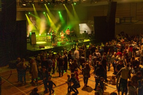 A band performs on stage with colorful stage lights in front of an audience in a concert hall.