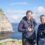 Two individuals stand on a cliffside wearing hoodies and binoculars around their necks, with a rocky coastline and ocean in the background.