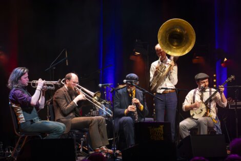 A jazz band performs live on stage, featuring musicians playing a trumpet, saxophone, trombone, and a sousaphone under colorful stage lighting.