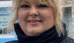 A woman with blonde hair and a black scarf smiles at the camera. She is standing outside in front of a stone building and a partially visible sign.