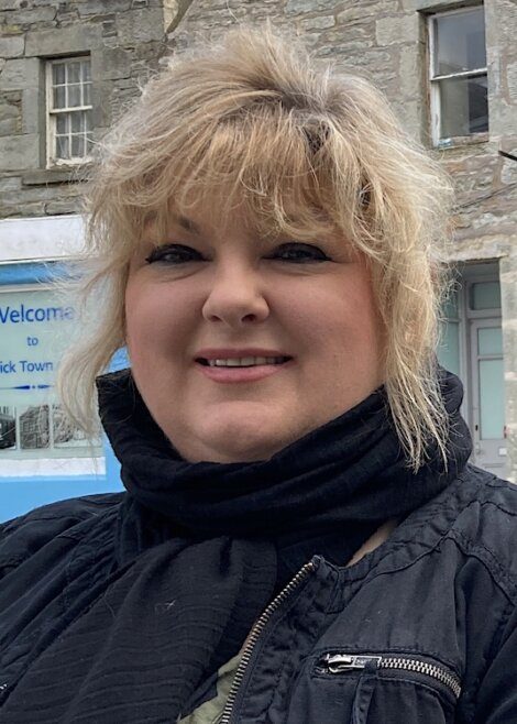 A woman with blonde hair and a black scarf smiles at the camera. She is standing outside in front of a stone building and a partially visible sign.