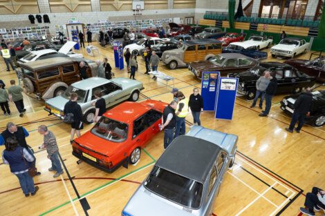 A gymnasium filled with different vintage and classic cars on display, with people walking around and observing.