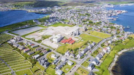 Aerial view of a coastal town with residential buildings, a cemetery, green spaces, and large construction areas near the shoreline and harbor.