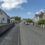 A small road on a sunny day with a 20 mph speed limit sign, a row of houses on both sides, and two cars parked along the curb.
